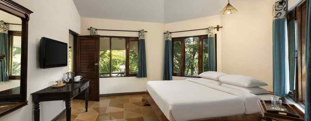 An image of a bedroom with double size bed, bedside table on either side, photos hanged on the wall and four windows from which natural lights are coming in - The Riverview Retreat,  Corbett
