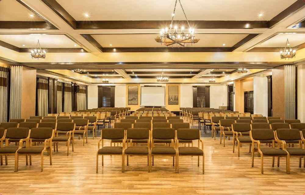 An image of a conference hall with a set of table and chairs in each corner and a projector screen at the front of the room - The Riverview Retreat, Corbett