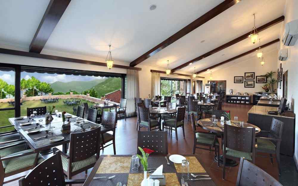An image of a Restaurant area with wooden chairs and tables which has plates and cutlery on top At baikunth Resort, Kasauli