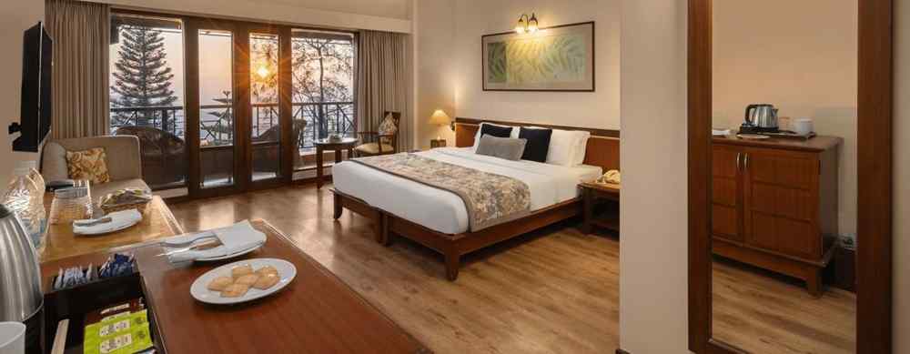 A wide view of a superior room showcasing a bed with side tables, sofas, a large wooden-framed mirror on the wall, and a balcony offering a sunny mountain view  - Baikunth resort, kasauli 