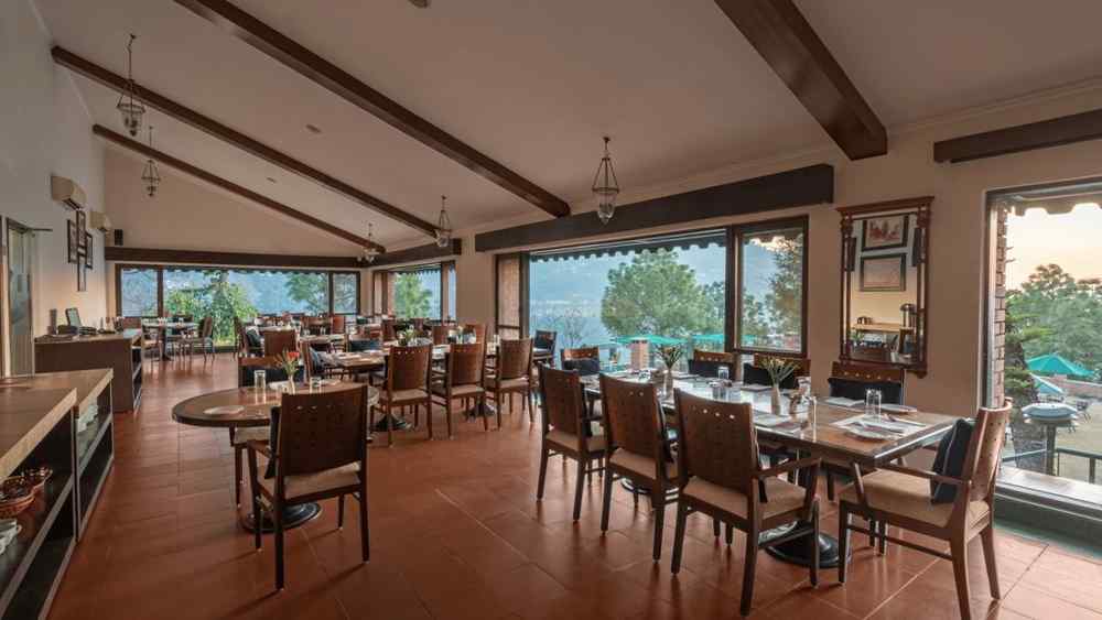 A restaurant with glass windows offering a beautiful mountain view, featuring dining tables arranged with chairs, a mirror on the wall, and stylish pendant lights overhead -  Baikunth resort, kasauli 