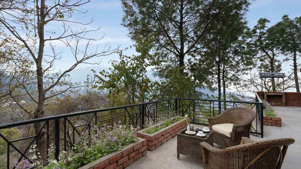 Two wooden chairs and a table set on an open balcony with a view of trees and mountains afar - Baikunth resort, kasauli 