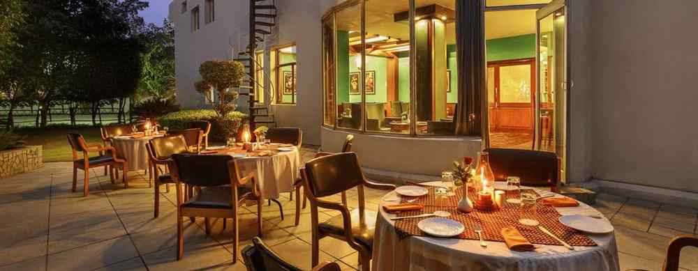 An outdoor dining setup with tables and chairs arranged outside, flanked by trees and neatly trimmed bushes