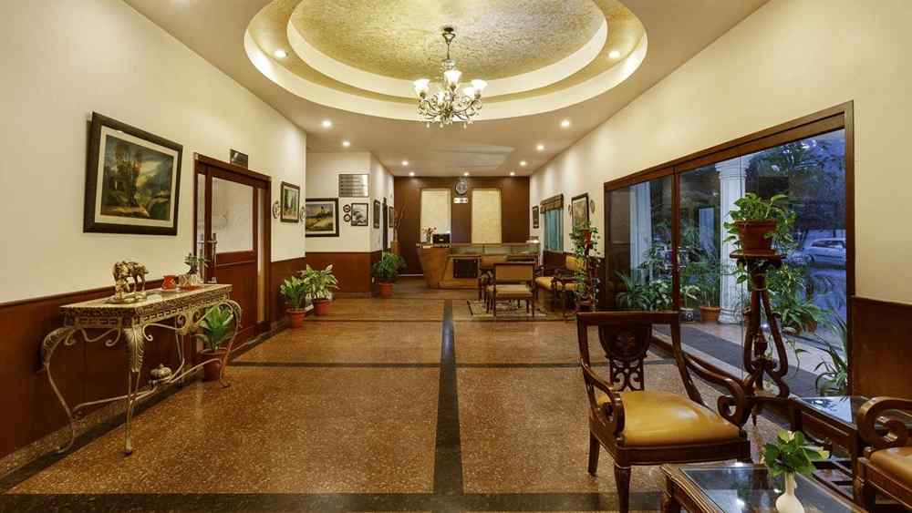 A corridor adorned with decorative frames, featuring high tables topped with flower vases and indoor plants, along with a few chairs arranged along the sides