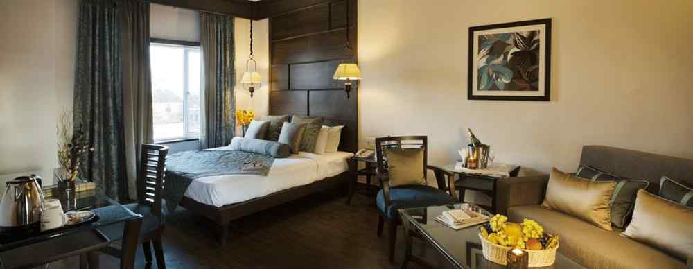A bedroom with a a bed placed on the corner beside a large window and a sofa and a table - The Manor, Kashipur