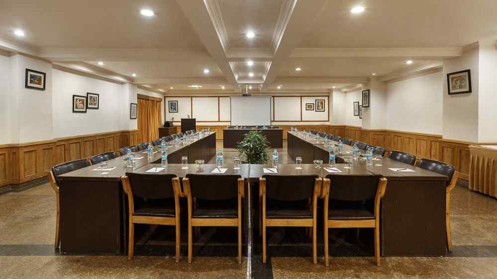 A spacious banquet hall features chairs arranged in a U-shape, illuminated by ceiling lights, and adorned with small photo frames on the walls
