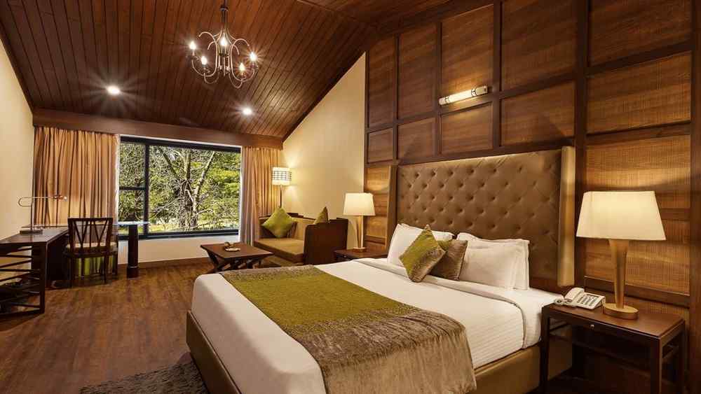 A bedroom with a large bed and a workdesk and chairs on the corner of the room, with large windows with trees visible - The Earl's Court, Nainital