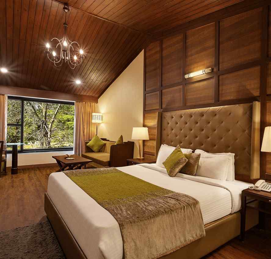 A bedroom with a large bed and a workdesk and chairs on the corner of the room, with large windows with trees visible - The Earl's Court, Nainital