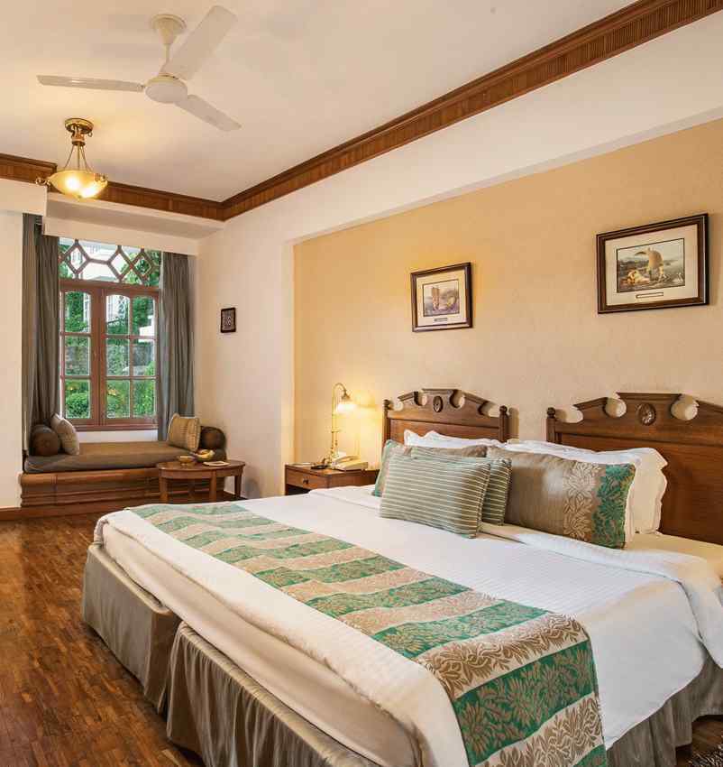 A well decorated room with a large bed, a shiny mirror alongside a make up table, with multiple wall decors and lights, portrayed while showcasing the lush outdoors through the doorhole- The Naini Retreat, Nainital.