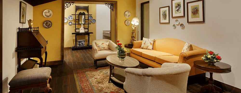 An image of the living area with Sofas that could seat 5 people, spaciously arranged furniture, multiple wall paintings, and a slanted wooden ceiling which accentuates the ambience.