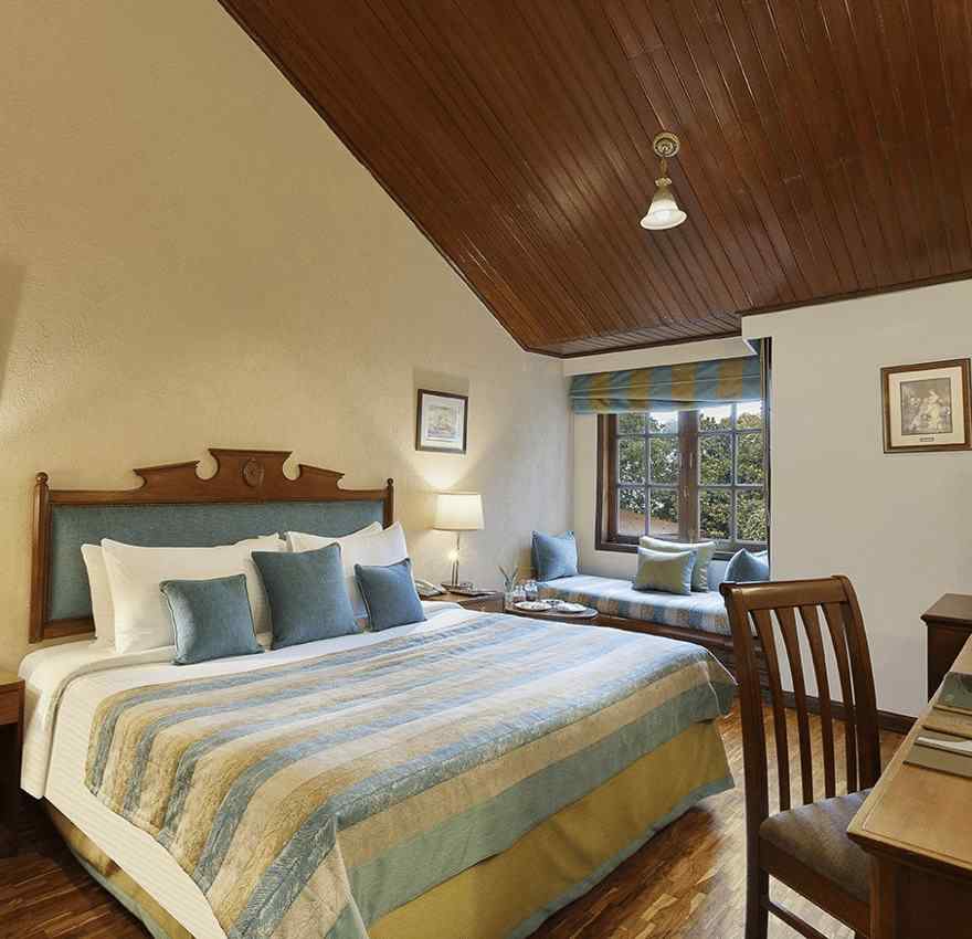 An image of a  cozy room in the naini retreat, nainital, with a king-size bed, wooden furniture, and a bay window overlooking a garden. The room features a ceiling fan, a television, and a small sitting area. 