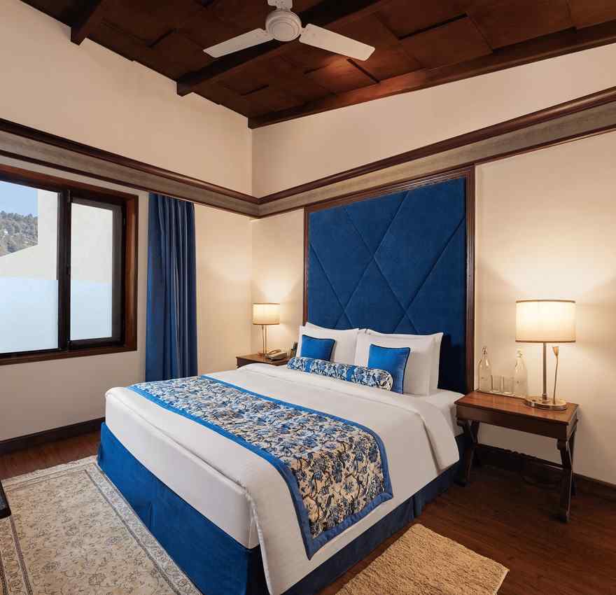 An image of a  cozy room in the naini retreat, nainital, with a king-size bed, wooden furniture, and a window. The room also features a ceiling fan.