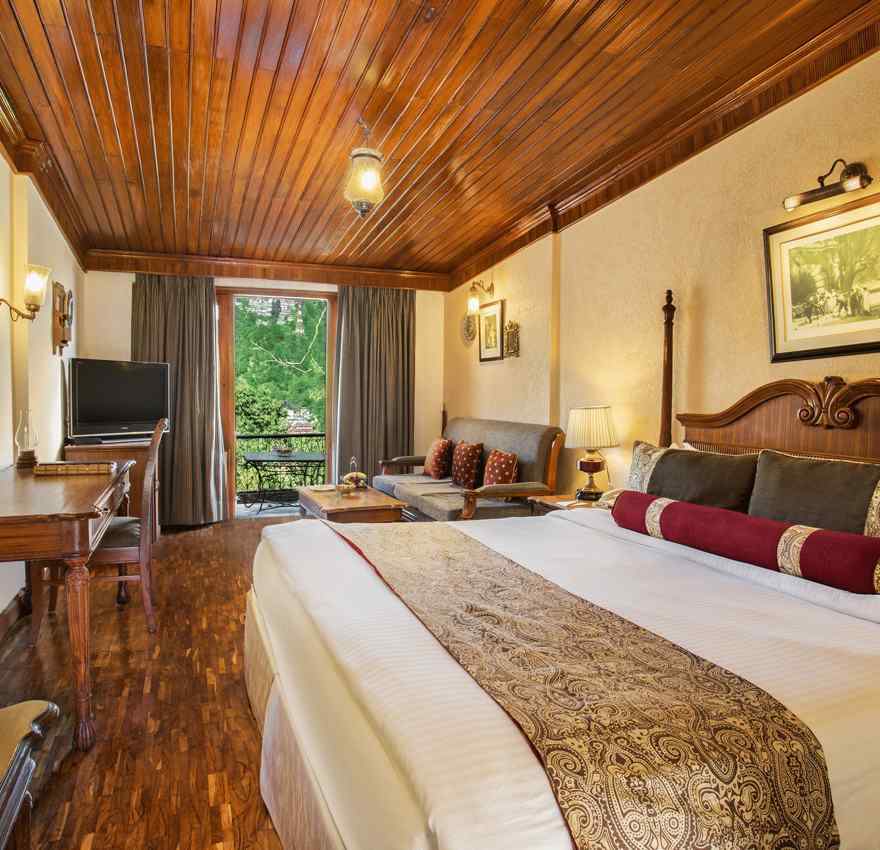 An image of a hill view room in naini retreat, nainital, with a king-size bed, wooden furniture, and a bay window overlooking a garden. The room features a ceiling fan, a television, and a small sitting area.