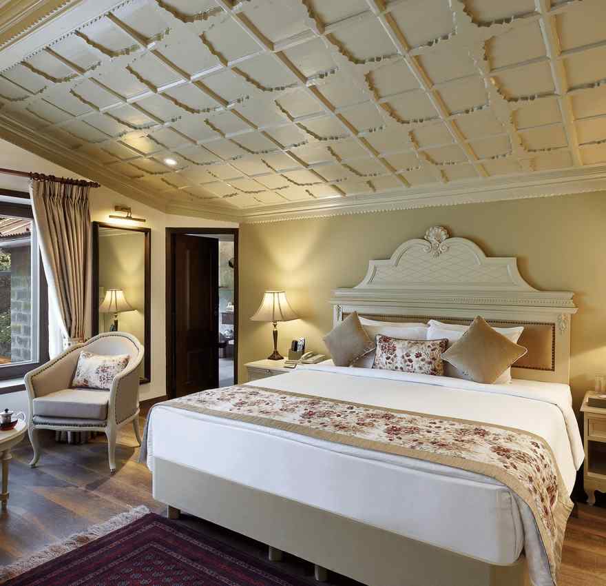 A full view of a room with a royal bed, chair, table and a huge glass window - the naini retreat, nainital.