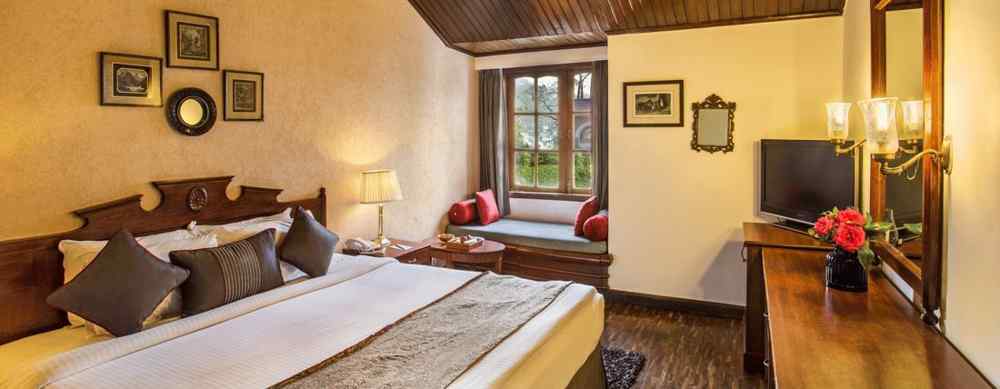 An image of a cozy room in naini retreat, nainital, with a king-size bed, wooden furniture, and a bay window overlooking a garden. The room features a ceiling fan, a television, and a small sitting area.