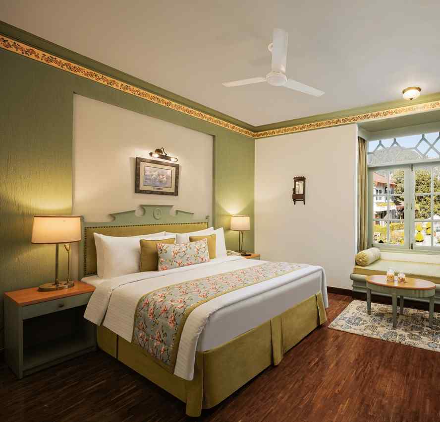 An image of a  cozy room in the naini retreat, nainital, with a king-size bed, wooden furniture, and a window. The room also features a ceiling fan, a TV and wooden flooring.