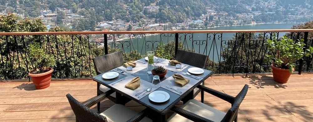 Image showing few chairs placed on the  sides of a square table, pot on either side of the frame, beautifully framed by the backdrop of the mountain ranges.