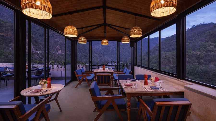 Indoor dining area at The Terrace, Aloha on the Ganges, Rishikesh, featuring wooden tables, pendant lighting, and floor to ceiling glass windows overlooking the hills.