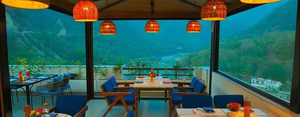 Riverside dining area at Aloha On The Ganges, Rishikesh, offering scenic views of the Ganges River and surrounding hills in a tranquil setting.