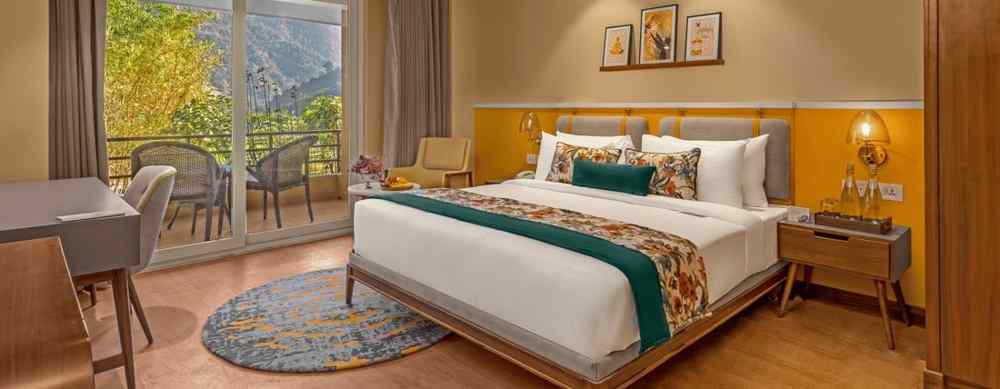 An image of a room featuring a large king-size bed with a headboard and comfortable bedding. A comfortable armchair sits by a large window offering breathtaking views. The room also includes a wooden desk with a chair, a television, and a ceiling fan. 