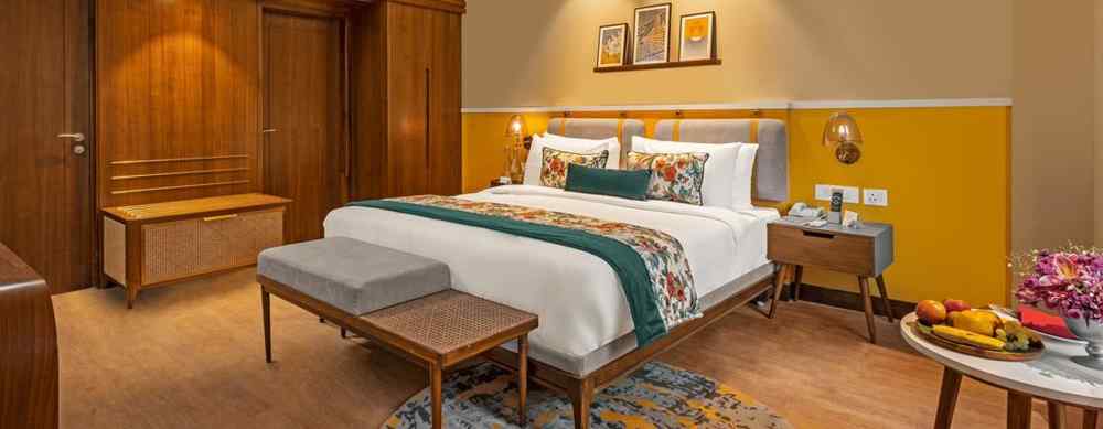 An image of a room featuring a king-size bed with a headboard and comfortable bedding. A bench sits at the foot of the bed, and a small table with a fruit platter is placed nearby.