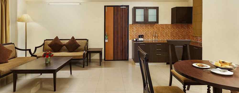 An image of a spacious and modern hotel suite. The living area features a comfortable sofa with throw pillows, a coffee table, and a floor lamp. A separate dining area