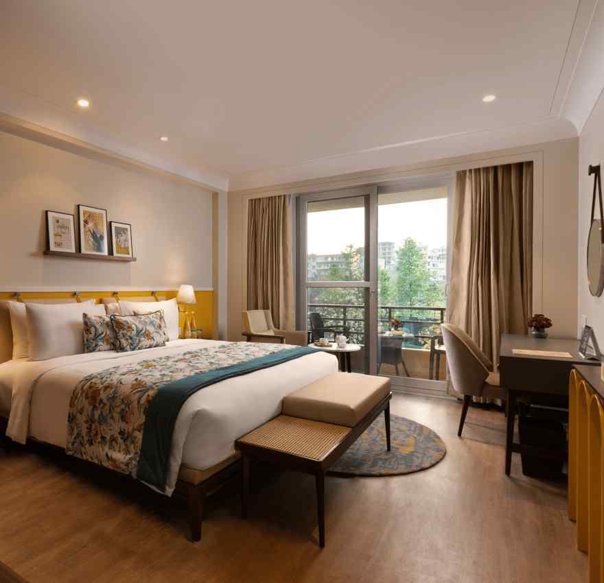 A spacious interior of room with a large bed, a sitting area with a sofa and a coffee table, and a desk. The room has large windows with curtains and a balcony overlooking a view.