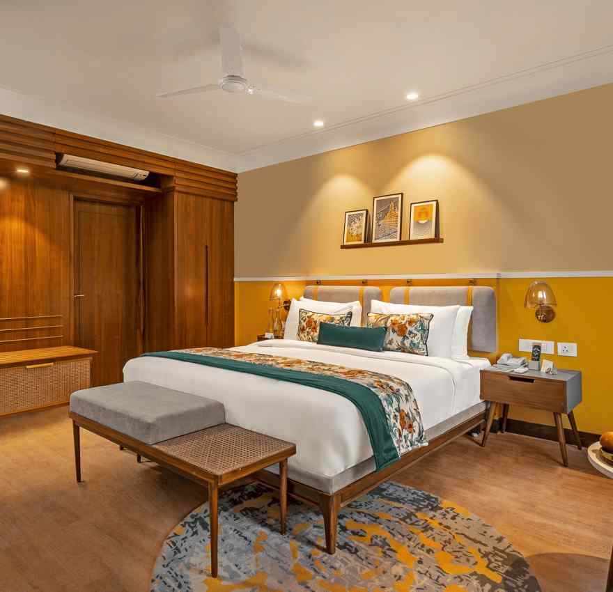 An image of a room featuring a king-size bed with a headboard and comfortable bedding. A bench sits at the foot of the bed, and a small table with a fruit platter is placed nearby.