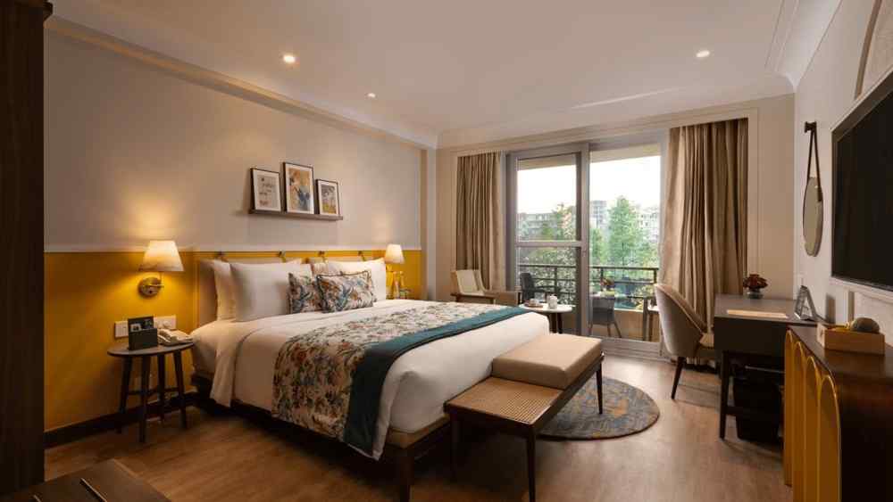 Elegant bedroom at Aloha on the Ganges Resort featuring a large bed with white linens, patterned pillows, warm lighting, balcony view, and cosy decor for a relaxing stay.