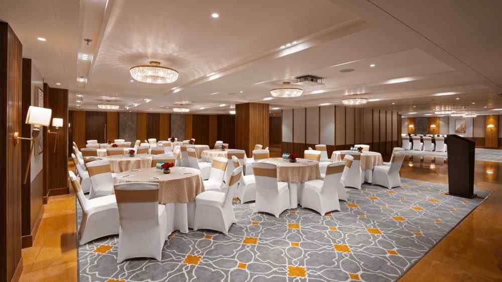 Spacious banquet hall at Aloha on the Ganges Resort with round tables, white chairs, patterned carpet, wood-paneled walls, chandeliers, and podium for events.
